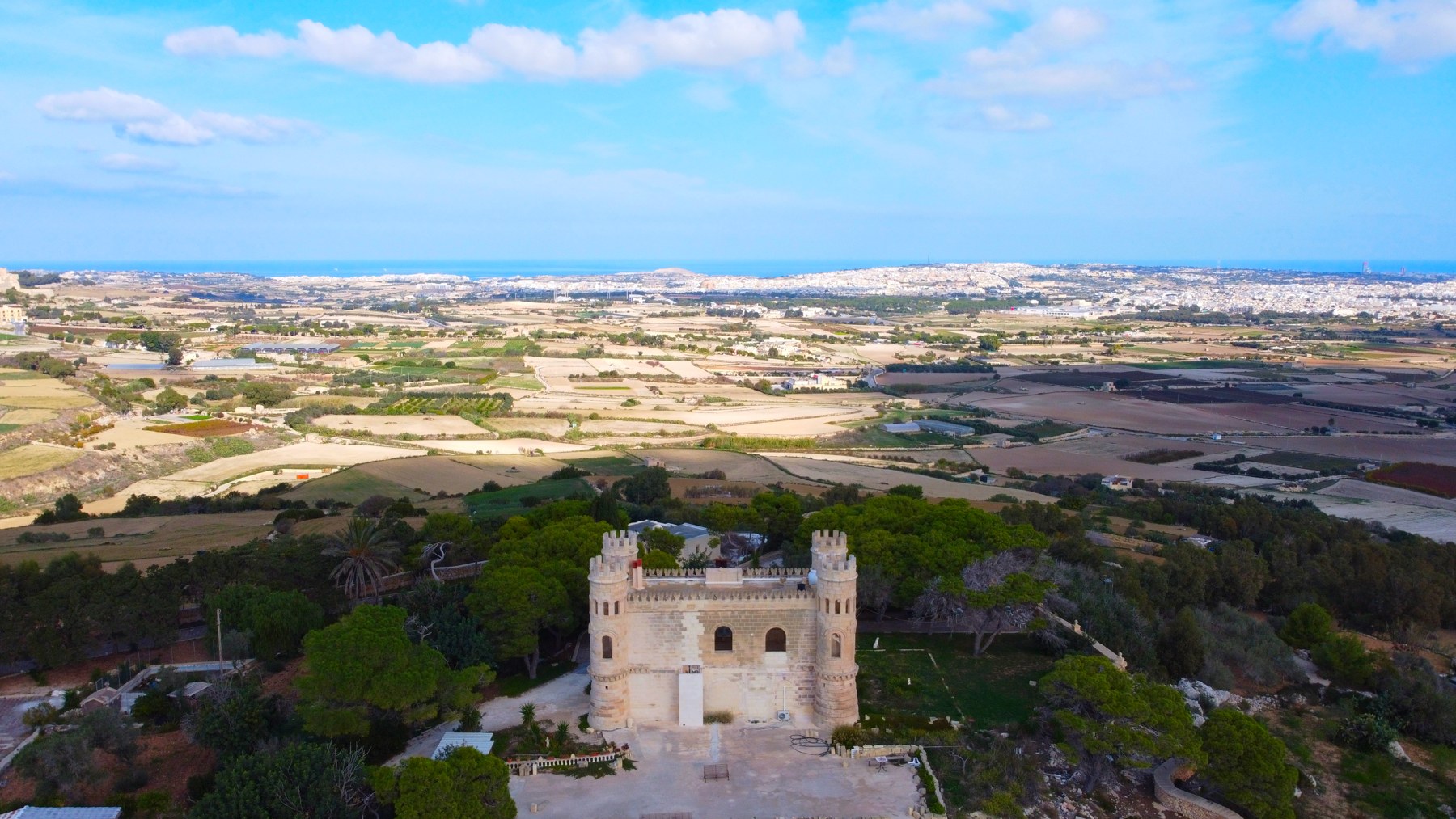 A turreted limestone building surrounded by trees and a broad panorama of Maltese fields