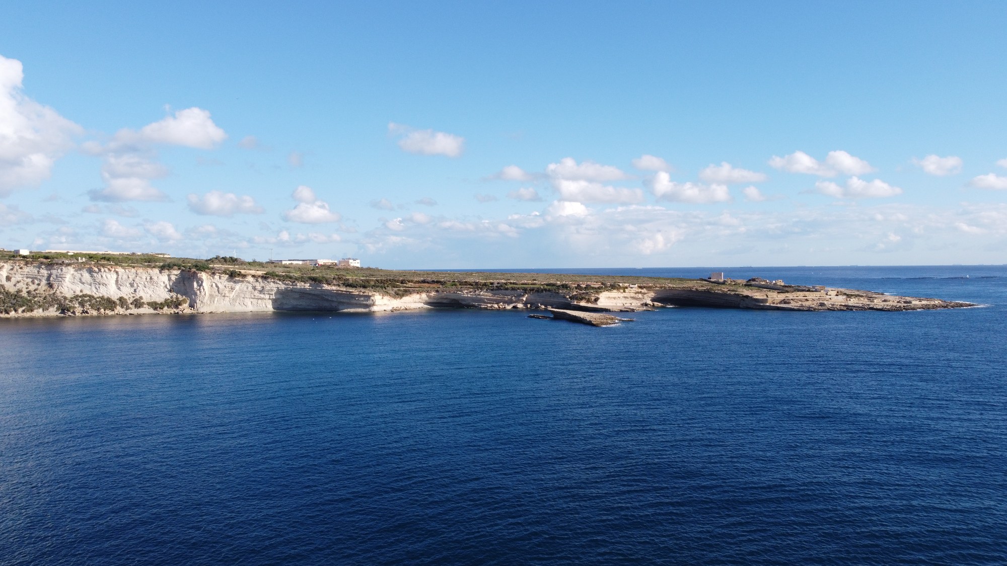 Wide aerial view across the blue water and low limestone coast of Delimara