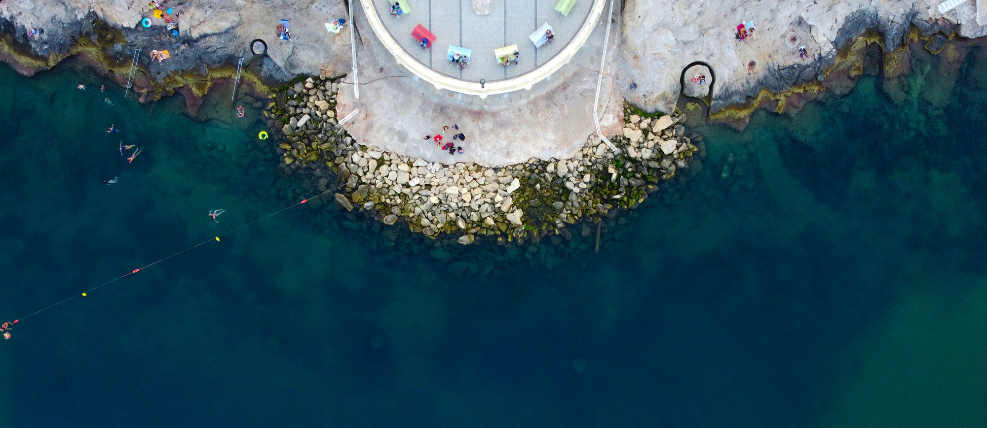 Top-down aerial view of swimmers beside a rocky seaside lido